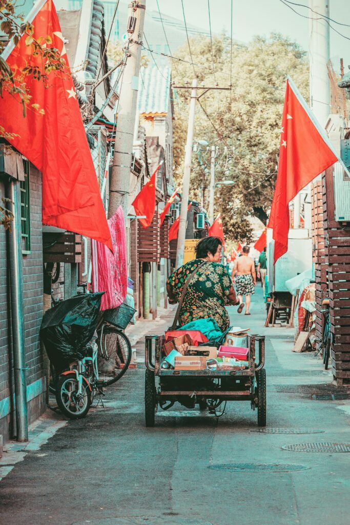 A narrow alley in Beijing featuring traditional architecture, red flags, and a man transporting goods on a cart.